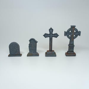 May include: Four miniature gray stone grave markers, two with crosses and two with rectangular headstones. One cross has a circle design and the words "R.I.P." on the base.