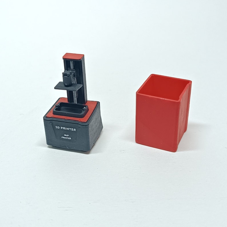 May include: A small black 3D printer with a red base and a red box. The printer has a label that says "3D PRINTER" and "DLP PRINTED".