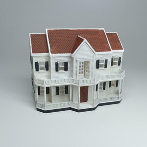 May include: A miniature white house with a brown roof, black shutters, and a porch. The model has a detailed design, including windows, doors, and railings, and is set against a plain background. This miniature house is a decorative item.
