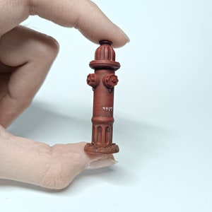 May include: A miniature red fire hydrant with a white number "1977" on the side. The hydrant has two black handles and a black base.