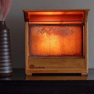 Könnte beinhalten: Ein hölzerner Honigextraktor mit beleuchtetem Wabeninneren. Der Extraktor hat eine aufklappbare Oberseite und den Text "BeeBreath" auf der Vorderseite. Eine braune Vase mit rosa und beigem Ziergras und eine Glasflasche mit Korken sind ebenfalls sichtbar.