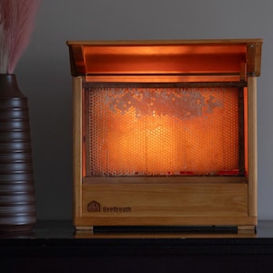 May include: A wooden BeeBreath honey extractor with a honeycomb interior illuminated by a warm light. A decorative brown vase with pink and white plumes and a small glass jar with a cork stopper are also present.