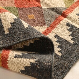 May include: A close-up of a woven rug with a geometric pattern in shades of brown, red, green, and white. The rug is folded over, showing the texture of the weave.