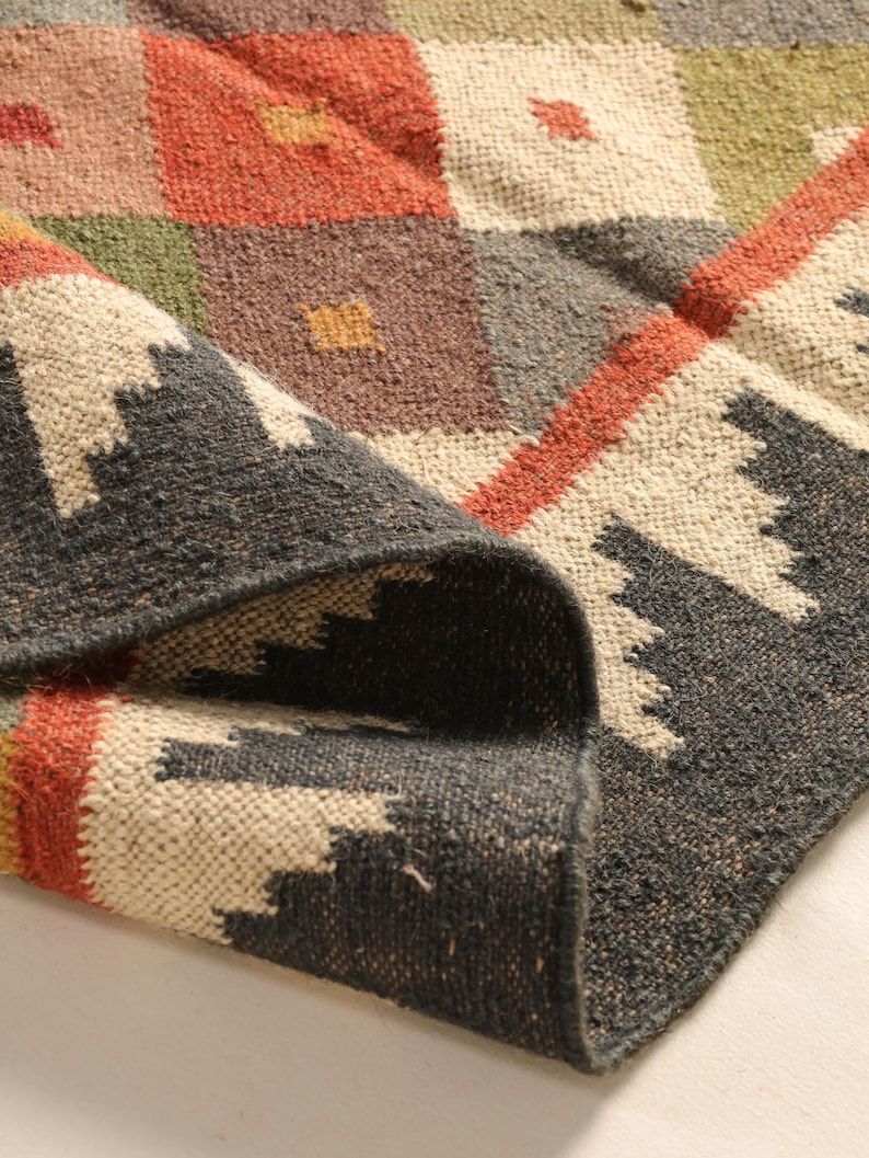 May include: A close-up of a woven rug with a geometric pattern in shades of brown, red, green, and white. The rug is folded over, showing the texture of the weave.