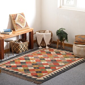 May include: A colourful, geometric patterned rug with black fringe. The rug is made of natural fibres and has a distressed, vintage look. It is laid out on a light grey floor in a room with a wooden bench, a woven basket, and a potted plant.