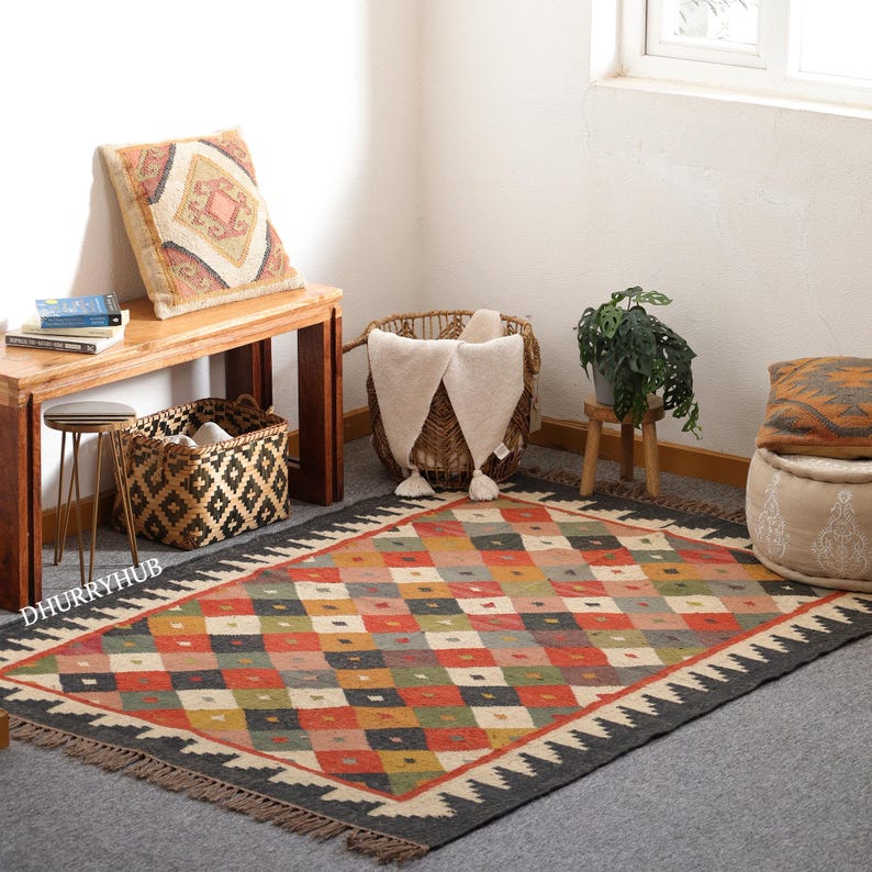 May include: A colourful, geometric patterned rug with black fringe. The rug is made of natural fibres and has a distressed, vintage look. It is laid out on a light grey floor in a room with a wooden bench, a woven basket, and a potted plant.