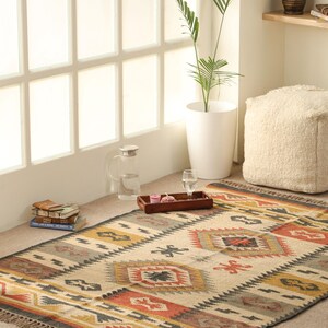 May include: A handwoven rug with a geometric pattern in shades of beige, brown, red, and yellow. The rug has a fringe border and is laid out on a light brown floor.