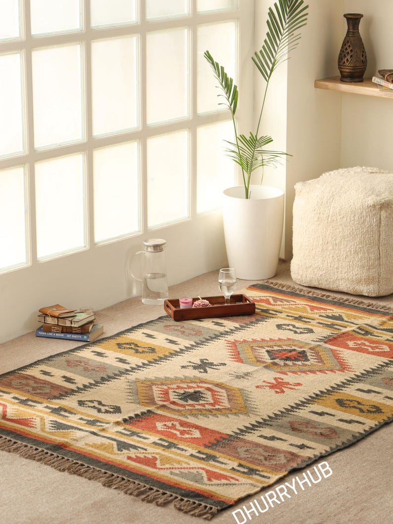 May include: A handwoven rug with a geometric pattern in shades of beige, brown, red, and yellow. The rug has a fringe border and is laid out on a light brown floor.