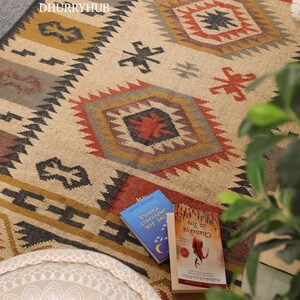 May include: A beige, brown, and black patterned rug with geometric designs. The rug is made of jute and wool and has a hand-woven texture. Two books are lying on the rug, one with the title "Give For The Things" and the other with the title "Chamatkara You Don't"