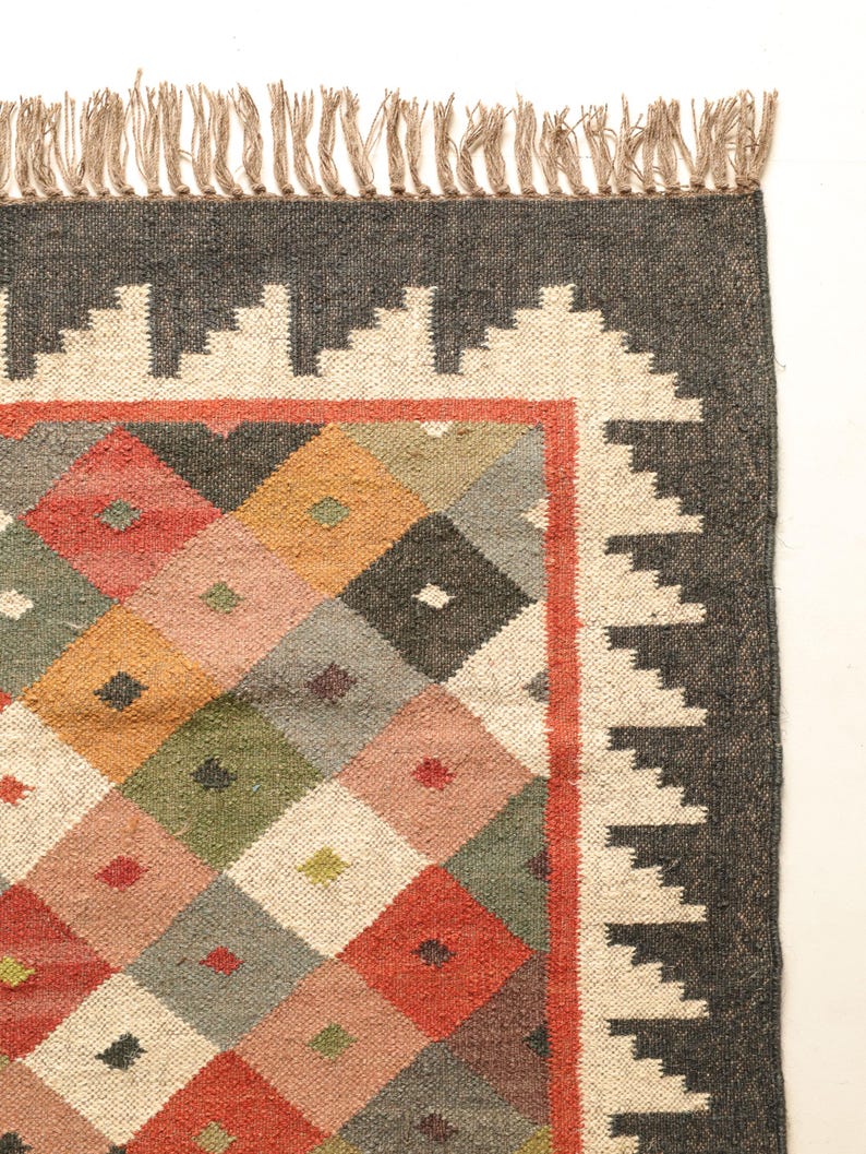 May include: A colourful rug with a geometric pattern of diamonds in shades of red, orange, green, blue, and white. The rug has a black border with a white fringe.