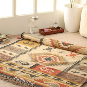 May include: A handwoven rug with a geometric pattern in shades of beige, brown, red, and yellow. The rug has fringe on all sides and the text "DHURRYHUB" is visible in the lower left corner.