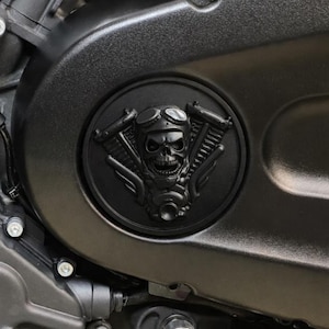 Könnte beinhalten: Schwarze Motorrad-Motorabdeckung mit einem kreisförmigen Emblem, das einen Schädel mit Brille zeigt, flankiert von Motorkomponenten. Das Emblem ist ein detailliertes, dreidimensionales Design. Abdeckung und Emblem sind mattschwarz.