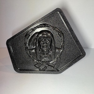 Könnte beinhalten: Dunkelgraue, rechteckige Metallschnalle mit einem detaillierten Relief des Gesichts eines indianischen Häuptlings in einem Kranz. Die Schnalle hat eine strukturierte Oberfläche und eine eckige Form.