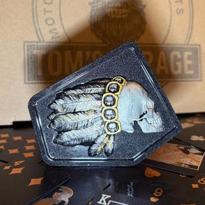 Könnte beinhalten: Ein schwarzes, rautenförmiges Emblem mit einem Totenkopf mit Federschmuck. Der Schädel ist silberfarben mit goldenen Akzenten, und die Federn sind in Grau und Gold detailliert. Das Emblem befindet sich vor einem Hintergrund aus Spielkarten.