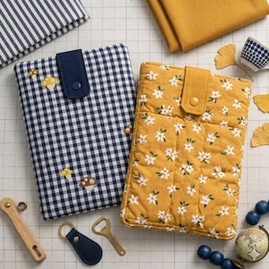 May include: Two fabric book covers. One is navy blue and white gingham with mushroom embroidery, the other is mustard yellow with white floral print. Both have a navy blue tab closure. Other items include a wooden bottle opener, a navy blue leather keychain, and a brass bottle opener.