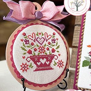 May include: A cross-stitch embroidery featuring a floral design in a basket, with pink and red accents. The embroidery is displayed on a stand and adorned with a pink bow. The word "February" is at the bottom. The text "Baskets of Blooms" is at the top.