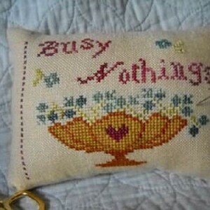 May include: A white needlepoint pillow with the text "Busy as a Bee" embroidered in red and yellow thread. The pillow features a basket of flowers in shades of yellow, orange, red, and blue.