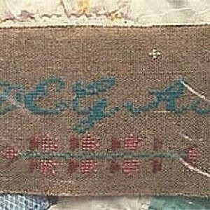 May include: A brown fabric sampler with the word "Beguiled" embroidered in blue thread. The sampler is decorated with a row of red and green flowers.