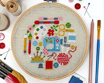 May include: A cross-stitch embroidery hoop featuring a sewing machine, scissors, and other craft supplies. The design is surrounded by a wooden hoop, with spools of thread, pencils, and a paintbrush nearby. The color palette includes blue, red, yellow, and pink.