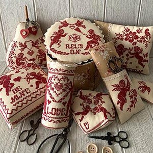 May include: A collection of handmade needlepoint pillows and a box featuring a Valentine's Day theme. The pillows are decorated with red and white embroidery of cherubs, hearts, and flowers. The box is decorated with a red and white embroidery of cherubs and the words "Really, Oh!"