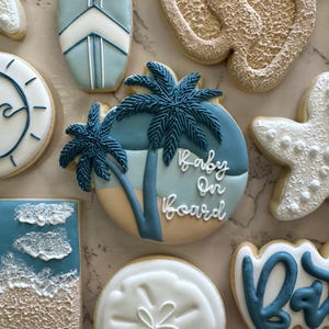 May include: Assortment of baby shower cookies with a beach theme. Cookies include a surfboard, footprints, a sun, a starfish, and a cookie with the text "Baby On Board" and palm trees. The cookies are decorated with blue, white, and tan icing.