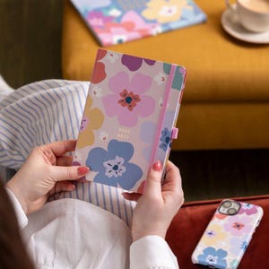 May include: A floral-patterned planner with the years 2026 and 2027 printed on the cover. The planner has a pink elastic band and a matching floral design. A phone case with a similar floral pattern is also visible.