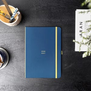 May include: A workspace scene featuring a blue 2026-2027 planner with a yellow elastic band, a cup of coffee, gold scissors, and a keyboard. The coffee cup has a blue and white design. The scissors are gold.