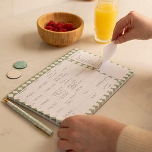 Pu&ograve; includere: Un blocco note verde e bianco con un motivo a rombi. Il blocco note ha una sezione per la colazione, il pranzo, la cena, gli spuntini e una lista della spesa. Il blocco note ha testo scritto a mano, tra cui "Colazione", "Pranzo", "Cena", "Spuntini", "Lista della spesa", "Questo", "Settimana", "Tacos", "Arrosto", "Limone", "Chia", "Beta", "Turinach", "Mat", "Bil", "Kjal", "Slinge", "2", "Trattare", "Lago", "Frutta", "Cereali", "Yogurt", "Arancia", "Succo", "Uova", "Latte", "Caff&egrave;", "T&egrave;", "Toast", "Avocado", "Insalata", "Zuppa", "Panino", "Pasta", "Pizza", "Pollo", "Pesce", "Verdure", "Frutta", "Dessert", "Spuntini", "Lista della spesa".
