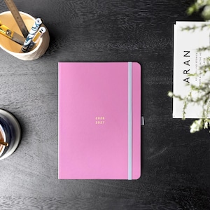 May include: A pink notebook with a light blue elastic band and the years 2026 and 2027 printed in gold. A cup of coffee, scissors, and a keyboard are on a black surface.