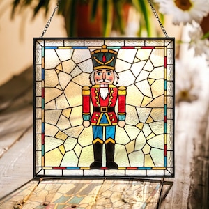 May include: A square stained glass suncatcher featuring a central nutcracker figure in a red, blue, and black uniform with a tall hat. The figure is set against a background of clear, textured glass pieces framed by a thin black metal border.