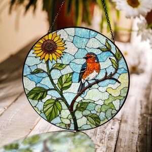 Pode incluir: Painel redondo de vitral com um girassol e um pássaro empoleirado num galho. O pássaro tem penas laranja, azuis e brancas. O fundo é um mosaico de vidro azul e branco, com folhas verdes.