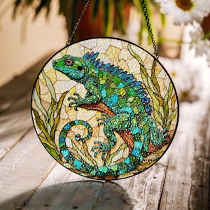 May include: A stained glass window hanging with a green and blue iguana lizard perched on a branch. The lizard has a curled tail and is surrounded by green leaves.