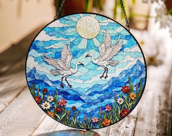 Può includere: Un acchiappasole in vetro colorato con due uccelli bianchi che volano sopra un paesaggio di montagna blu e bianco con un sole giallo e fiori in basso.