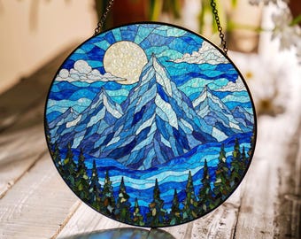 Può includere: Una finestra in vetro colorato appesa con una scena di montagna blu e bianca con una luna piena nel cielo. Le montagne sono in primo piano con una foresta di alberi alla base.