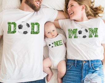 May include: A family of three wearing white t-shirts with a soccer ball theme. The father's shirt says "DOD", the mother's shirt says "MOM", and the baby's shirt says "ONE".