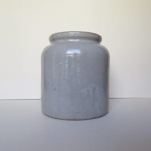 May include: A cylindrical ceramic crock with a smooth, glossy light grey glaze. The vessel features a thick, rounded rim at the top and a flat base. The surface shows subtle variations in the glaze texture, indicating a handcrafted finish.