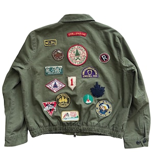 Peut inclure: Une veste vert olive ornée de divers écussons brodés au dos, incluant des textes comme "Camp Falls Scout Landing", "RL-1967", "Polo RL Est. 1967 Fishing" et "North and Fly Fishing Club". La veste présente un col classique.