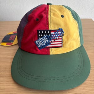 Può includere: Un cappellino da baseball multicolore con pannelli rossi, gialli, verdi e viola. La parte anteriore presenta una toppa ricamata di una bandiera americana con il testo SNOW BEACH RALPH LAUREN in lettere blu. Un'etichetta abbinata è visibile sul lato.