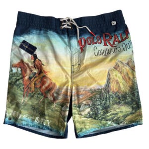 May include: Blue and yellow Polo Ralph Lauren swim shorts with a landscape design. The shorts feature a Native American on horseback, mountains, and the text "Colorado Out." They have a drawstring closure and a small tag.