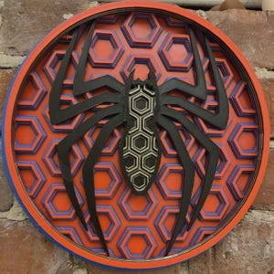 Puede incluir: Una silueta de araña negra sobre un fondo hexagonal rojo, azul y plateado en capas. La araña está centrada en la obra de arte de pared circular de madera.
