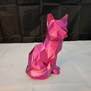 May include: A pink geometric cat figurine made of plastic. The cat is sitting with its tail curled around its legs.