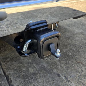 May include: A black metal trailer hitch with a silver pin and a silver loop. The hitch is attached to a vehicle.