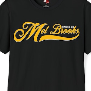May include: Black t-shirt with the text "Mel Brooks" in a yellow script font with a white outline, and the words "RAISED ON" above the name. The shirt is a crew neck style.