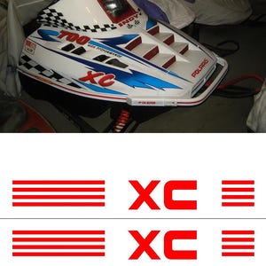 May include: A white and blue Polaris snowmobile with red detailing. The snowmobile features the text "700 XC HIGH PERFORMANCE" and "POLARIS". The design incorporates a chequered flag pattern and the letters "XC" in red.