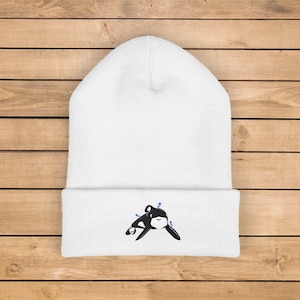 Op de afbeelding: Witte beanie met een zwart-wit orka-ontwerp geborduurd op de voorkant. De orka heeft blauwe accenten. De hoed is gemaakt van zacht, gebreid materiaal.
