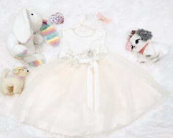 May include: A white tulle dress with a satin ribbon and lace detailing. The dress is on a white fluffy surface with stuffed animals.
