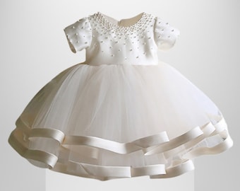 May include: A white tulle dress with a satin bodice and pearl embellishments. The dress has a round neckline and short sleeves. The skirt is layered with tulle and has a satin ribbon trim.