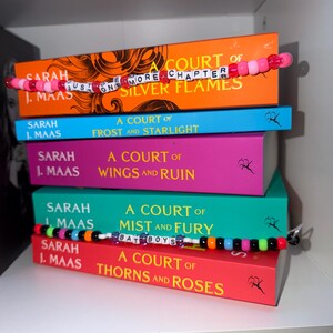 Puede incluir: Una pila de cinco libros de Sarah J. Maas con lomos coloridos. Dos hilos de cuentas están colocados sobre los libros. El hilo superior tiene cuentas rosas y letras blancas que forman JUST ONE MORE CHAPTER. El hilo inferior tiene cuentas multicolores y letras blancas que forman BAT BOYS.