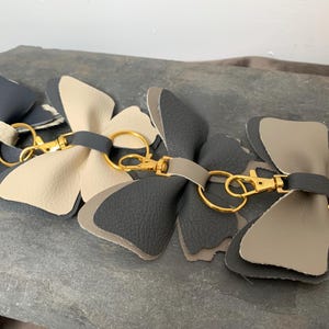Op de afbeelding: Drie lederen vlinder sleutelhangers met goudkleurige metalen ringen en een karabijnhaak. De vlinders zijn in de kleuren beige, grijs en zwart.