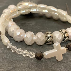 Op de afbeelding: Twee witte kralen armbanden met een zilveren bedel en een witte kruisbedel. De armband onderaan heeft heldere kristallen kralen en zwarte kralen.